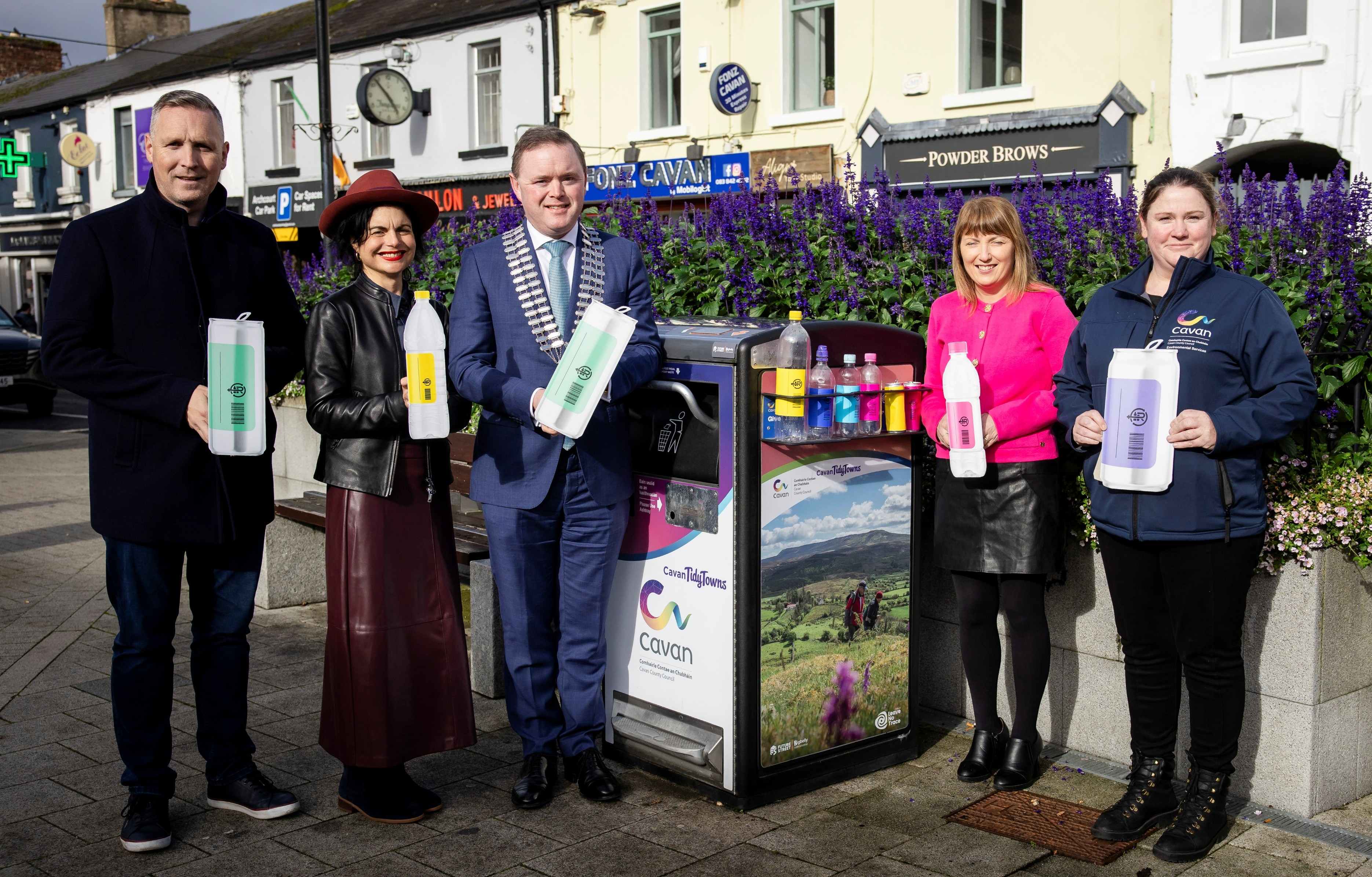 Five people standing on a town street holding oversized Re-turn branded drink containers in front of a Reverse Vending Machine. The center person wears mayoral chains of office, flanked by community representatives. The RVM displays Cavan Tidy Towns branding and contains visible plastic bottles inside. Purple flowering plants line the street planters behind the group, with traditional shopfronts including Powder Brows visible in the background.