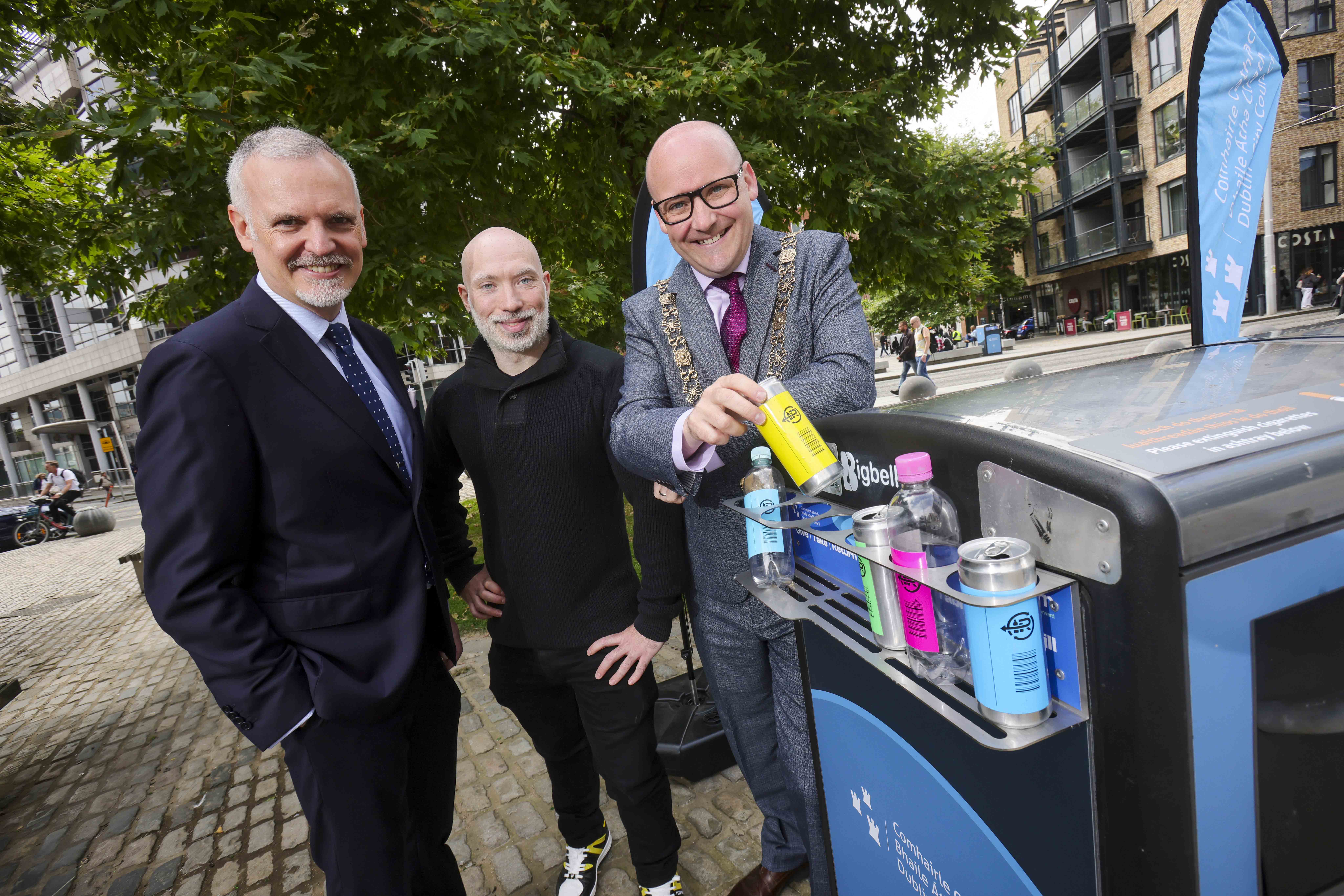 Three people beside a recycling bin on a cobblestone street; one is placing a yellow can into the bin, which contains various cans and bottles. Trees, buildings, and pedestrians are visible in the background.