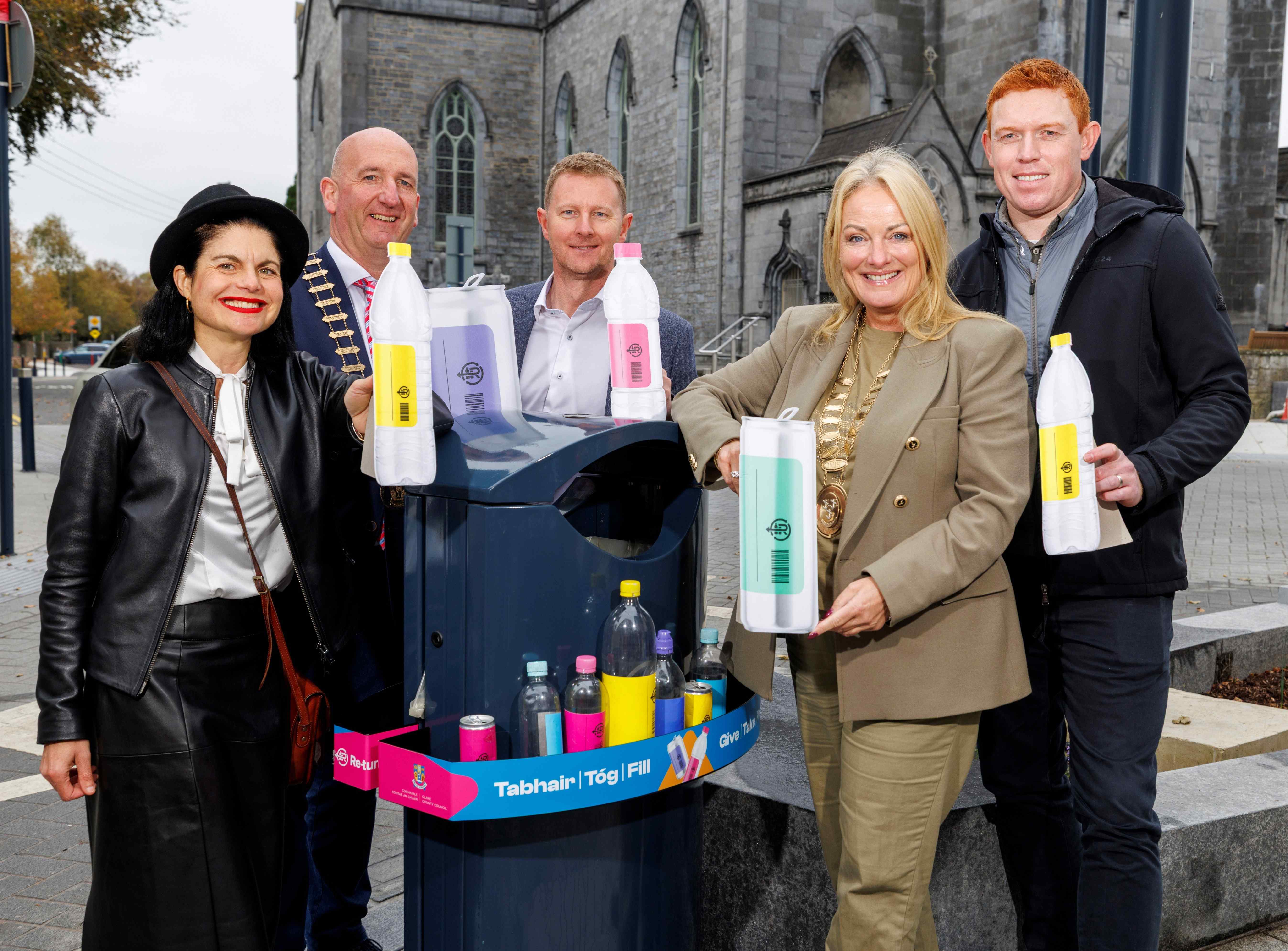 Five people standing around a recycling bin wrapped in bilingual signage, each holding a plastic bottle, with a historic stone building in the background. The image promotes environmental awareness and public recycling.