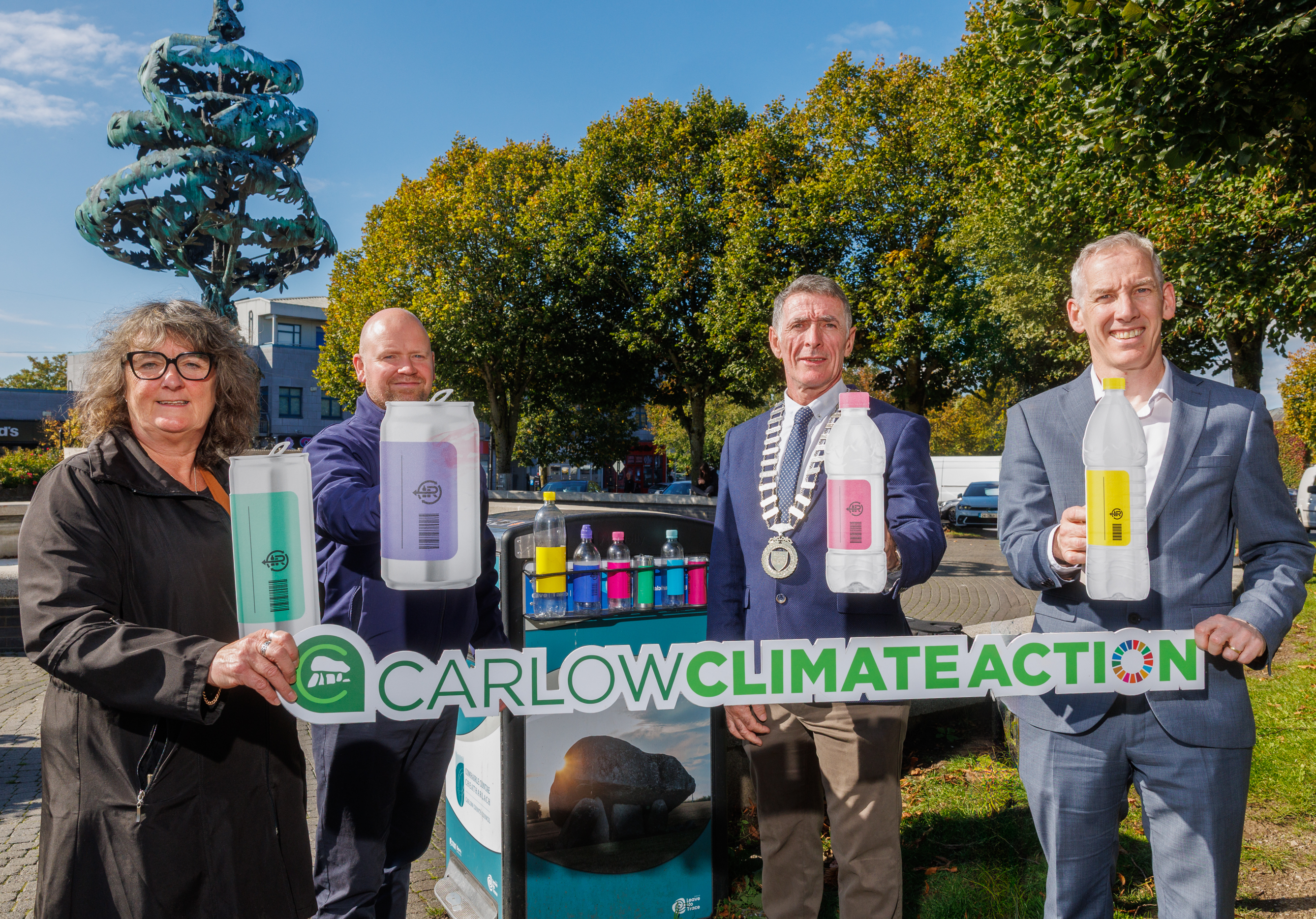 Four individuals standing outdoors in front of a 'Carlow Climate Action' sign, holding large plastic bottles filled with colored liquids. They are dressed in formal and semi-formal attire, with trees, a sculpture, and buildings visible in the background, suggesting a community event focused on environmental awareness.