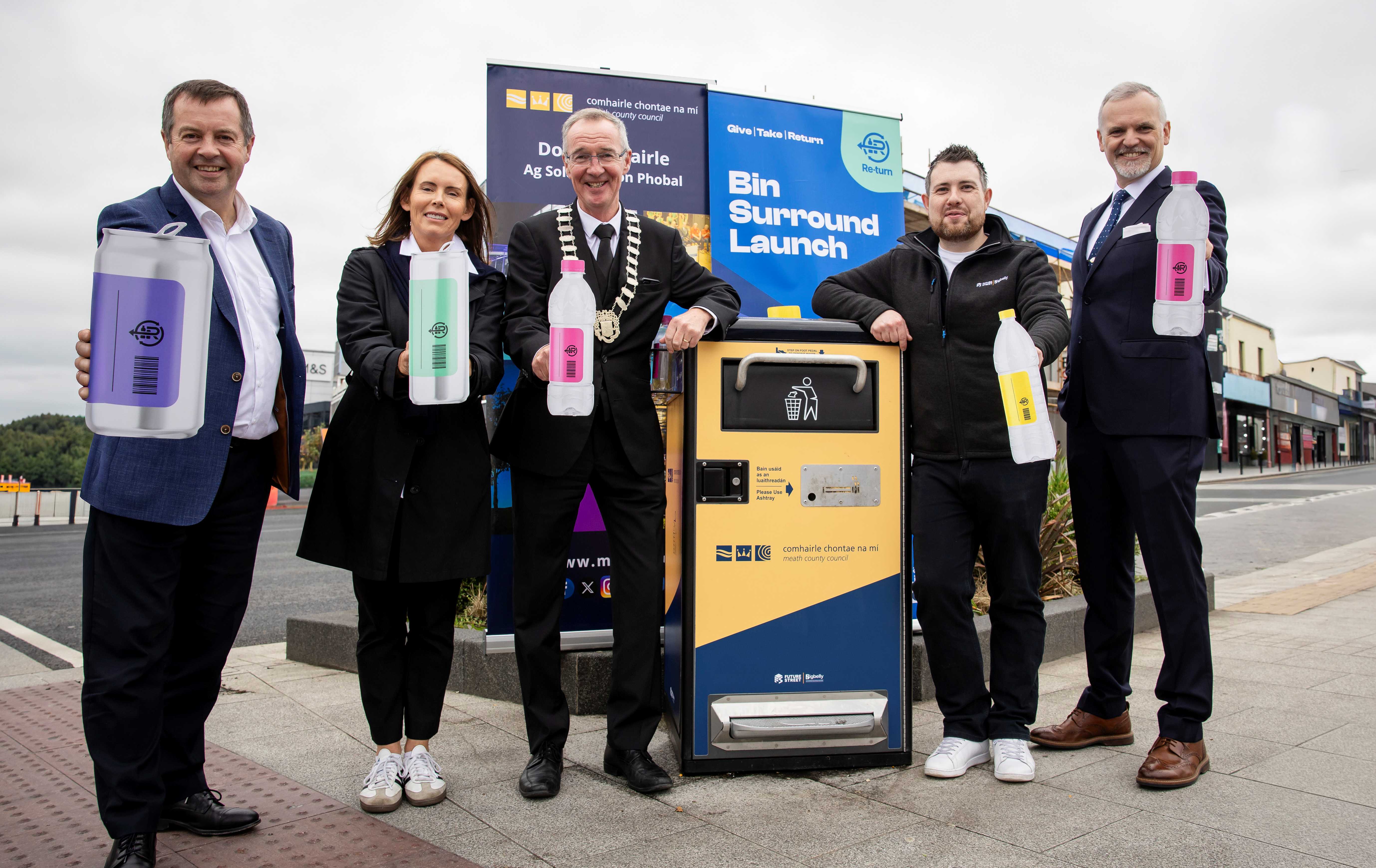 Five individuals standing beside a public bin labeled 'Bin Surround Launch,' each holding a large plastic bottle. The group is dressed in formal and semi-formal attire, with an urban street and buildings in the background, suggesting a recycling or waste management initiative.