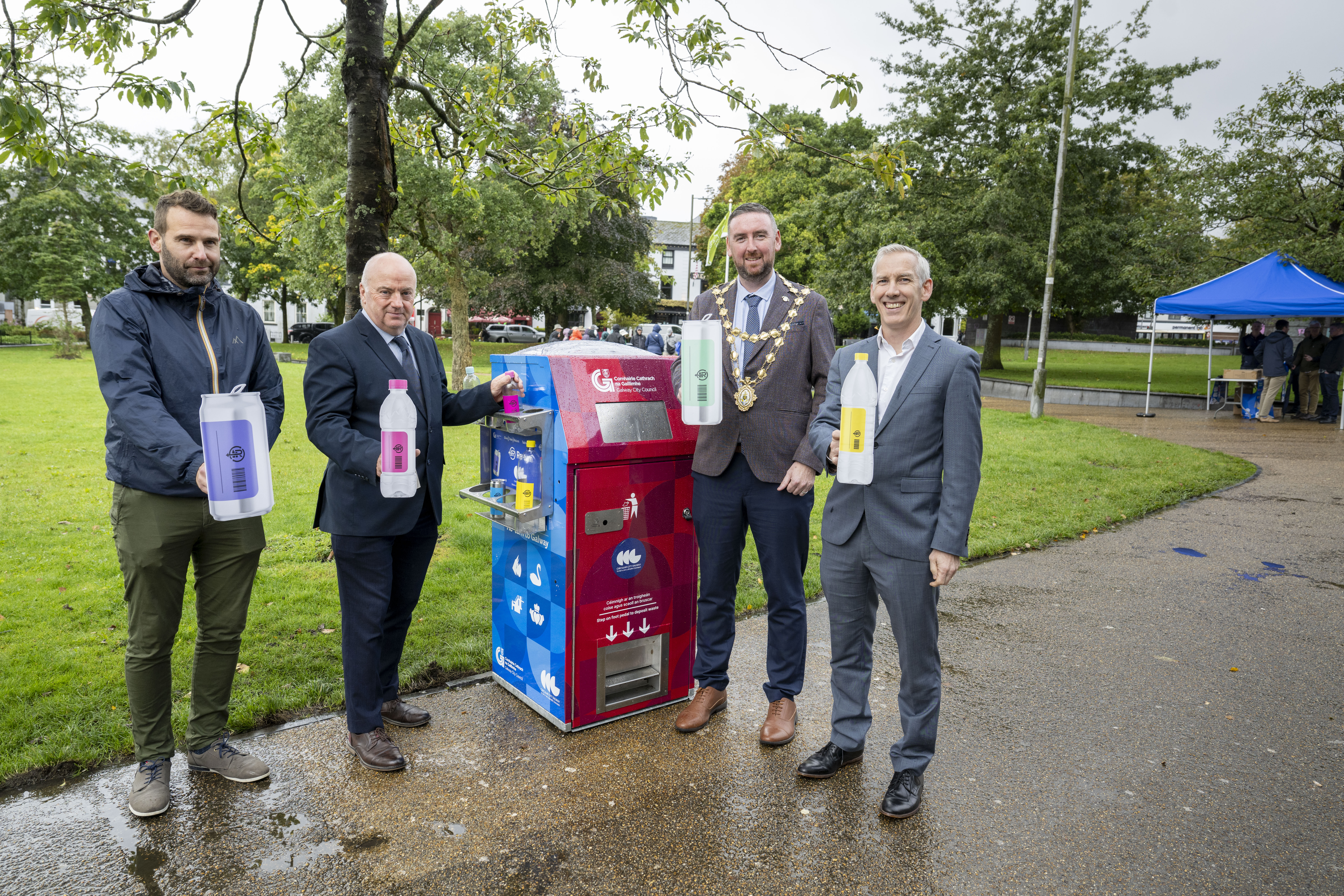 Four people standing in a park holding large plastic bottles near a colourful recycling machine, with trees, grass, and a blue tent in the background, likely at an environmental awareness event.